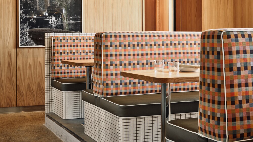 The interior of Paperboy South features booths made out of white tile with patchwork-patterned cushions. The walls are wooden and have a black and white photograph hanging.