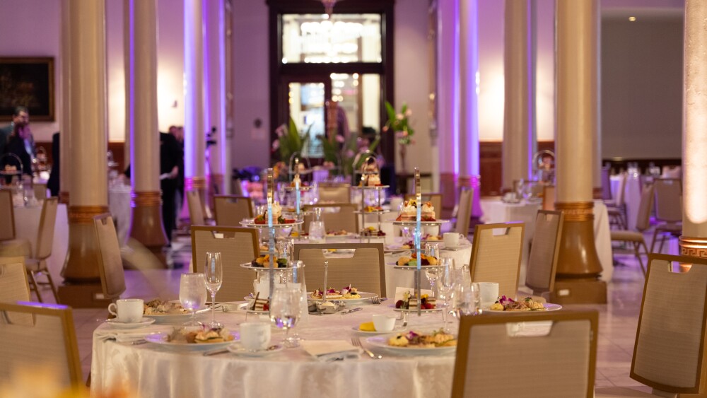 A banquet hall set for an event with round tables covered in white linens, each adorned with tiered trays of food, glassware, and floral garnishes, surrounded by beige chairs and tall columns under warm lighting with purple uplighting accents.
