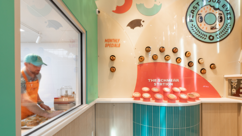 A wall displaying all the shmears, bagels, and monthly specials the business offers with a look at the rolling window on the left.