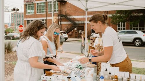 Two women speak to a small business vendor on a street.
