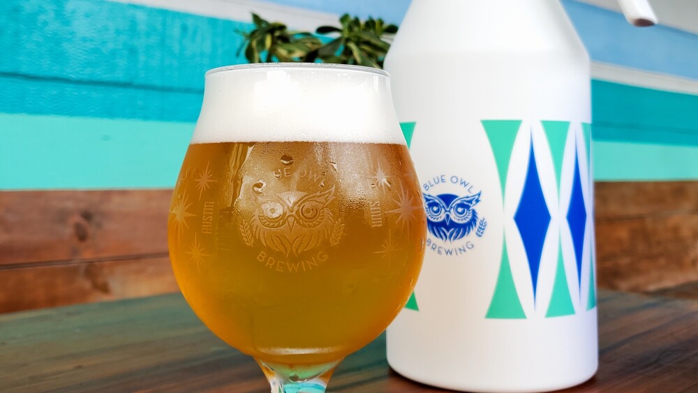 A large beer in a round glass sits on a mahogany table, in front of a blue, white, and teal flash, and a blue wall.