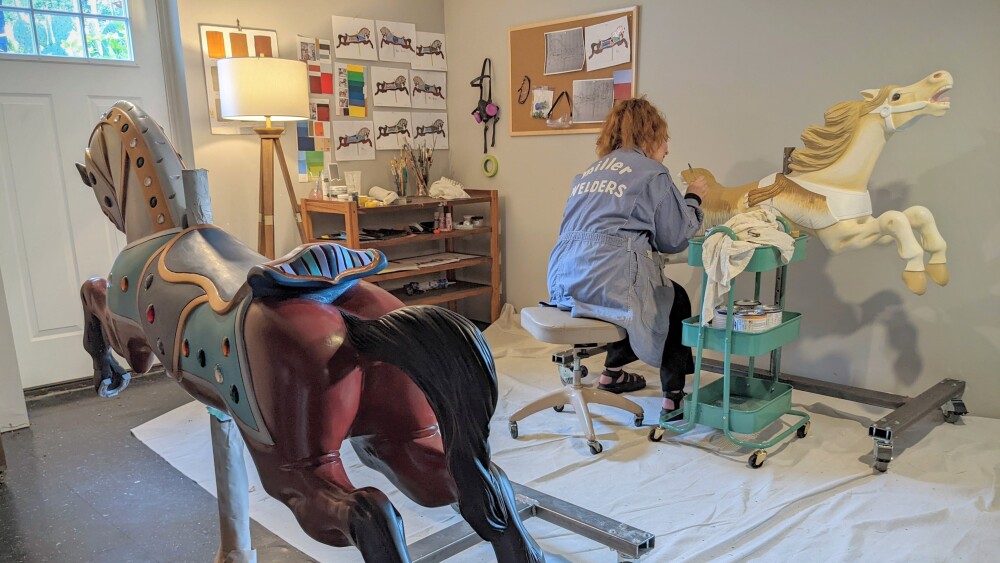 Two carousel horses are in a room. A person leans over one of them with a paintbrush.