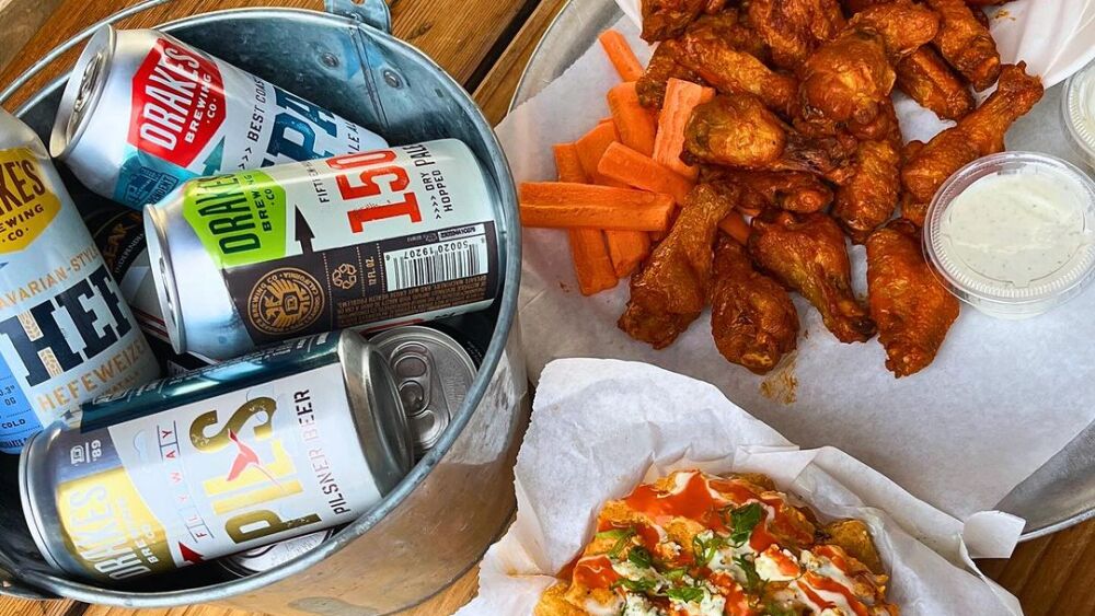 A bucket of beers, a pile of chicken wings, and a pizza atop a wooden table.