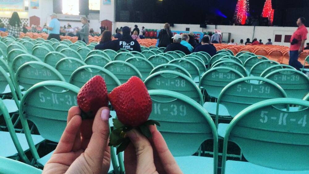 Two people hold strawberries from their seats in front of the Florida Strawberry Festival stage. The seats surrounding them are teal, and a few other audience members sit closer to the stage.