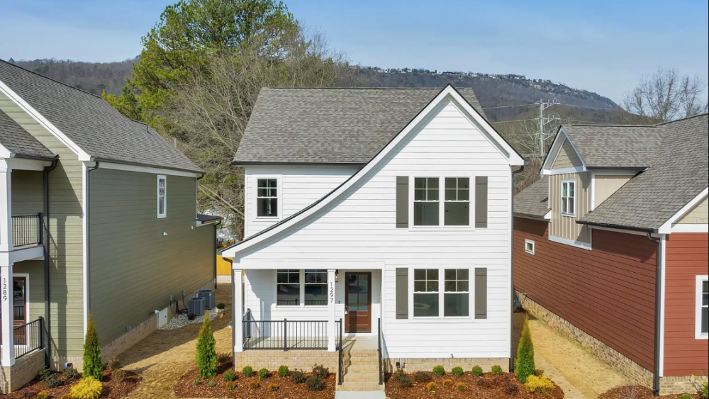 Modern white house with a front porch, flanked by similar homes. Gray roof, manicured garden, and a clear blue sky create a welcoming atmosphere.