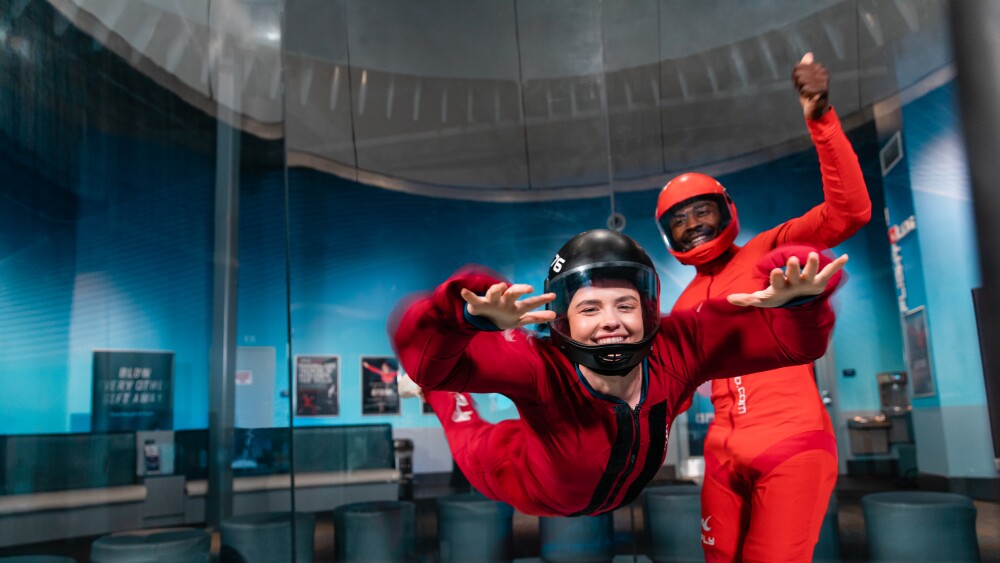 A person in a red jumpsuit and helmet hovers in the air and a spotter stands behind inside a glass-walled circular chamber.