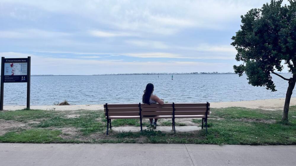 A woman sitting on a bench at Bayside Park overlooking San Diego Bay and the city skyline in the distance.