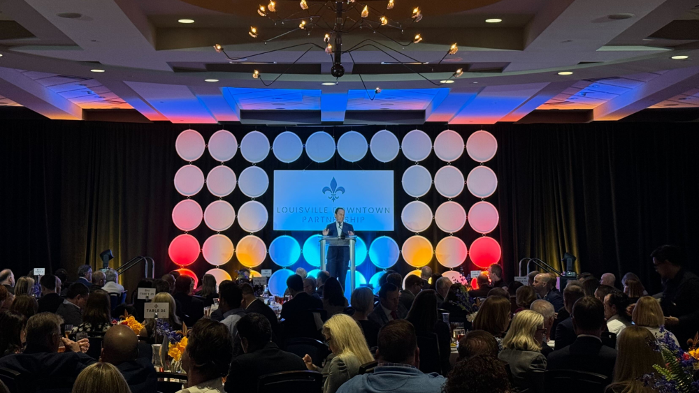 Mayor Craig Greenberg stands at a podium in front of a display meant to invoke stacked bourbon barrels + a sign reading Louisville Downtown Partnership while a room of people sit at round dining tables.