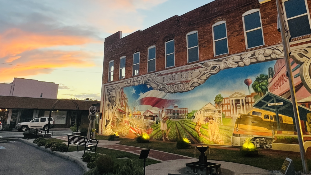 The back of a downtown building painted with a mural depicting Plant City's history. The sun is setting in the background behind the building. Several benches face the mural, and a walking path in front of them includes a fountain and a sculpture.