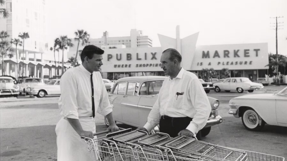 Publix founder George Jenkins talks to an employee pushing shopping carts outside of a Publix Super Market. The black and white photo is taken c. the 1950s or 1960s, with vintage cars around them in the parking lot.