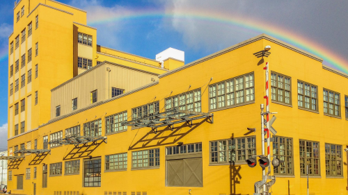 A yellow warehouse next to train tracks with a rainbow arching over its roof.