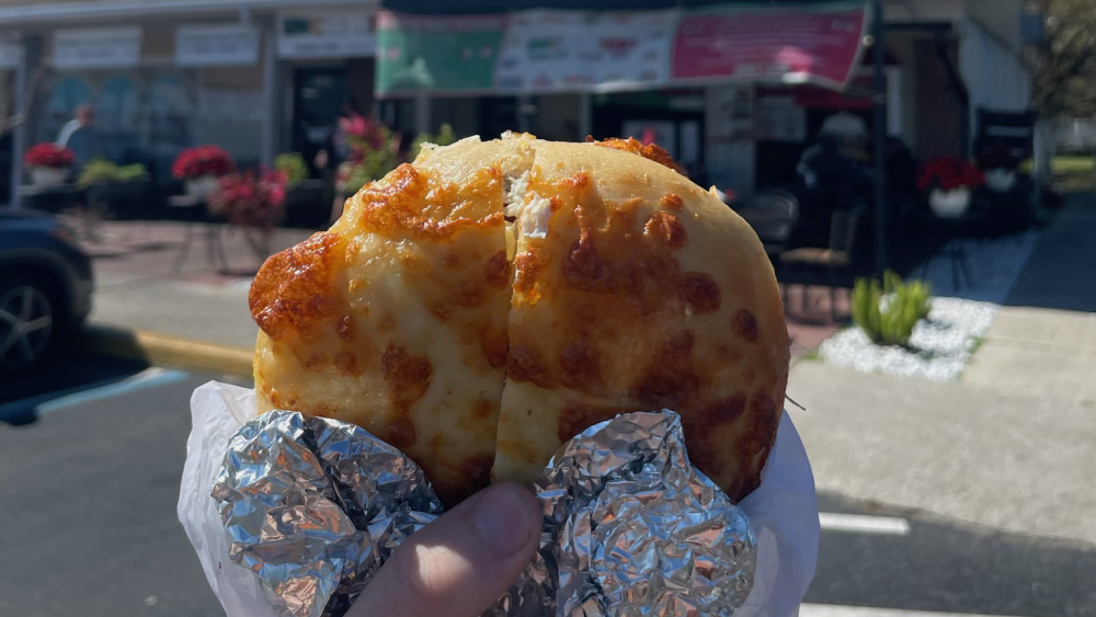 A hand holds a bagel sandwich in front of Uncle Nick's Deli in Lakeland. The cheese bagel is wrapped in aluminum foil. The deli is in a small shopping center and has a few tables and chairs by the front entrance.