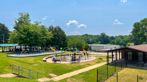 Green grass with payground, splash pad on concrete, a picnic shelter in the distance and a brick building with a black fence on the right.