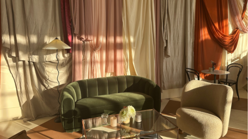 A green velvet love seat next to an armless white sherpa chair surrounding a gold and glass coffee table with a tapestry on the wall behind them.