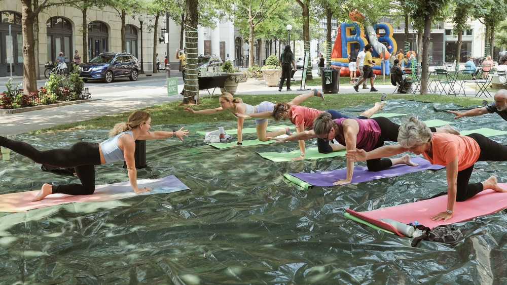 A small group of people do a yoga pose on their knees on their yoga mats, which sit on a tarp in Church Street Park.