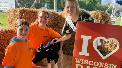 Two girls with a baby cow sit with Alice in Dairyland who holds a I Love Wisconsin Dairy Farmers sign.png