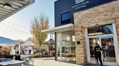 The outside of a coffee shop on a sunny day. Several chairs and tables are sitting outside in front of a building that says "Night Swim Coffee."