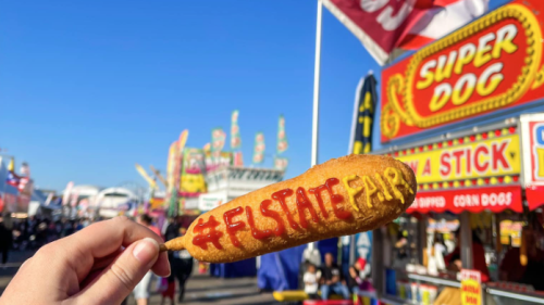 A corndog at the Florida State Fair. The words "#FL State Fair" are written on the corndog in ketchup and mustart. There are blue skies in the background above a variety of food trucks and stalls.