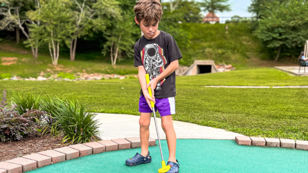 A young boy holding a yellow putter standing on a green at the McPherson Park mini golf course.