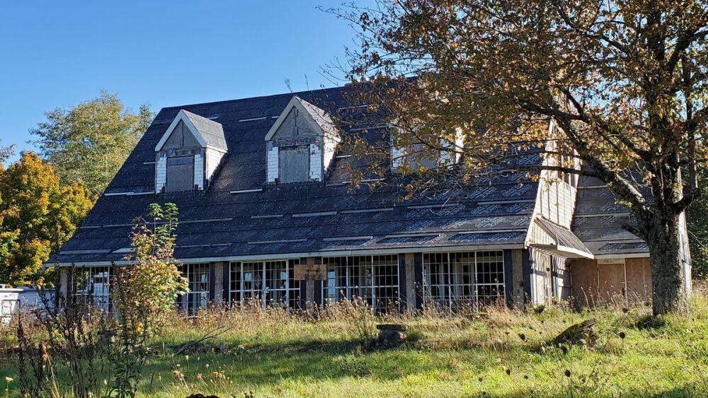 A neglected former inn with boarded-up windows is surrounded by grass and trees.