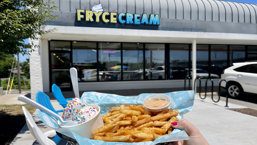 A hand holding up a basket of fries and a cup of soft serve in front of a business with a sign reading "Fryce Cream."