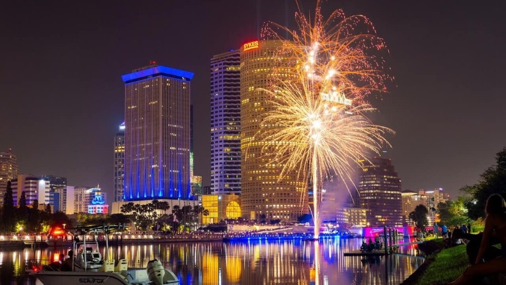 Orange and yellow fireworks burst over the water before two high rises, near a green shore with trees. Colorful lights from the city are reflected in the water.