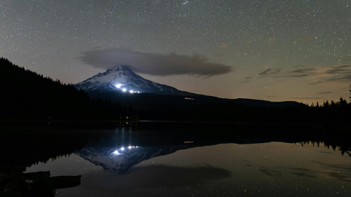 Mount Hood rises above a still lake at night, reflecting the stars above it. A cloud sits just atop its peak and lights from ski resorts on its slope shine bright.