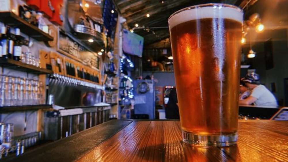 A glass of beer on a wooden bar at Swan Brewing. In the background are beer taps and customers.