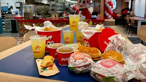 Taken inside the new Rainier Valley Jollibee location, this photo shows four chicken sandwiches, a few of the peach mango pies, two Chickenjoy buckets, and a tower of Pineapple Quencher sodas. A statue of Jollibee's bee mascot is gesturing enthusiastically at the menu behind the counter in the distance.