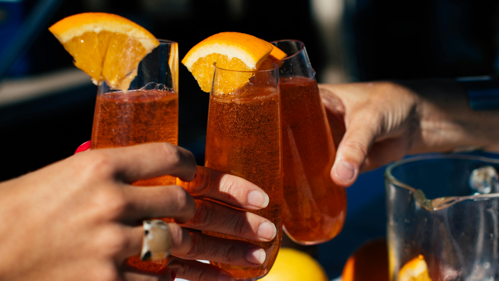 Three hands are holding bright orange, fizzy drinks rimmed with an orange wedge and toasting; a table covered in grapefruit and oranges serves as a backdrop.