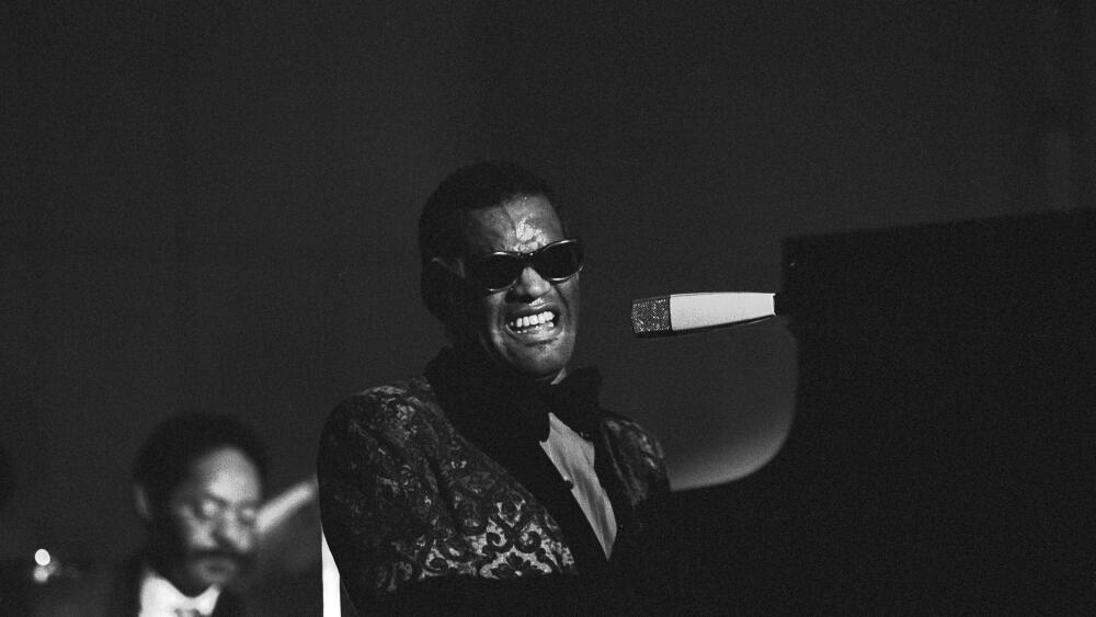 A black and white photo of Ray Charles playing the piano and singing. His iconic sunglasses are on and he's wearing a patterned suit which singing into a microphone.