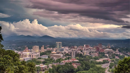 Asheville skyline from a distance with white and purple clouds above it.