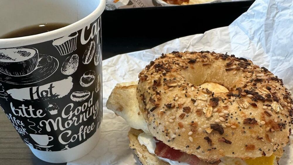 A breakfast sandwich sits on a table next to a cup of hot black coffee. The sandwich is made on an everything bagel with egg and bacon. Another meal sits further away on the table.