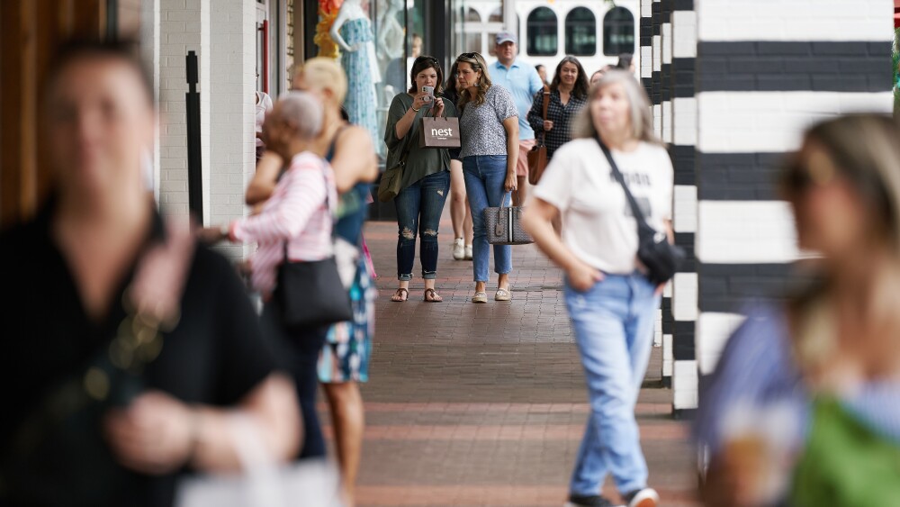 A crowd on the sidewalk outside of an open air shopping mall, with the camera focused on two women holding shopping bags