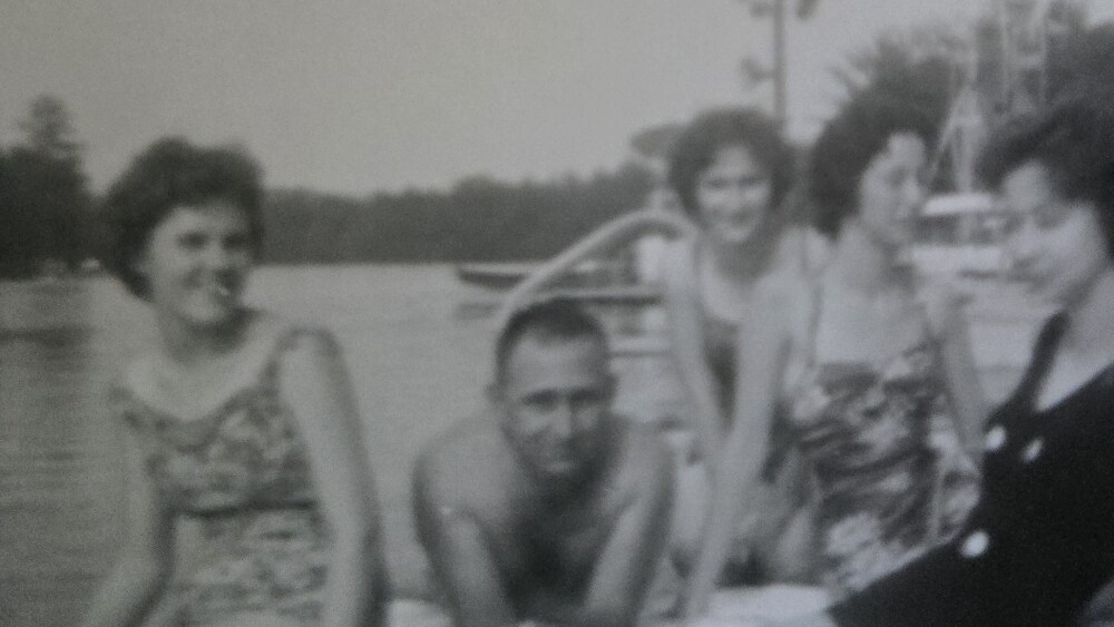 Vintage black and white photo of four women and a man in swimsuits on a dock by a lake. They are smiling, creating a relaxed and joyful atmosphere.