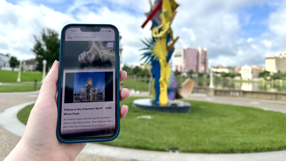 A hand holding a smartphone showing a sculpture Lakeland's downtown public art trail. The real sculpture is seen in the background.