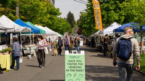 A block that goes through the Columbia City Farmers Market in Seattle, WA, with a green sign highlighting vendors and people browsing tented booths