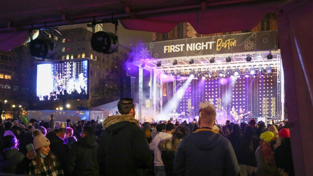 A crowd in winter clothes gathers in front of a stage with a sign that reads "First Night Boston"