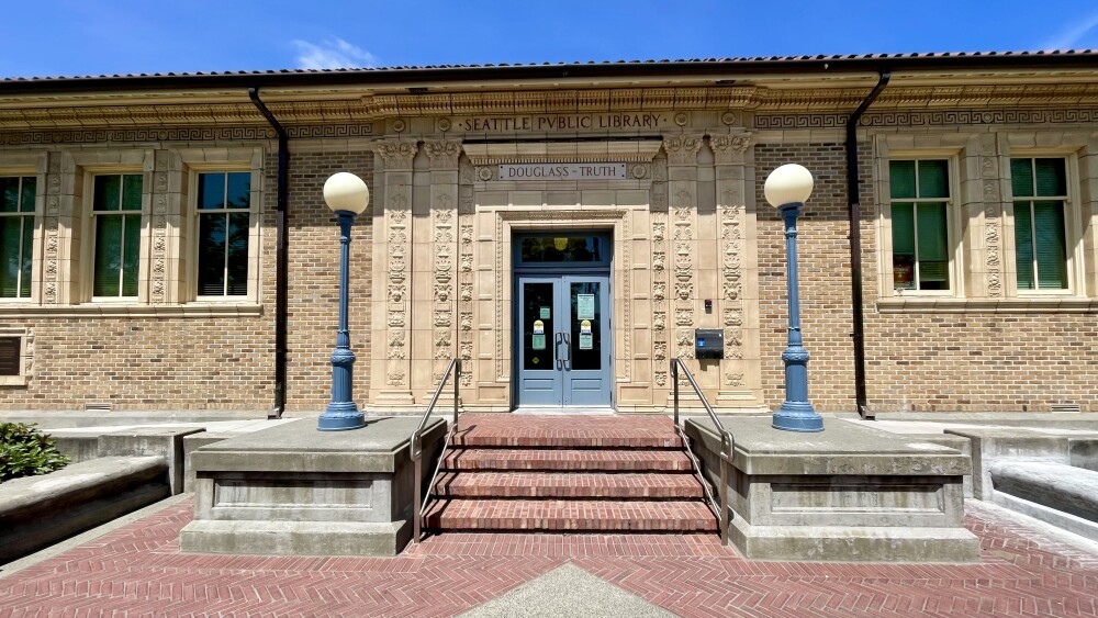 The yellow brick of Douglass Truth Library is decorated with stone blocks, carved into which is the name of the library. Lamp posts with circular tops rest on either side of the entry way's steps leading to the front's double doors.