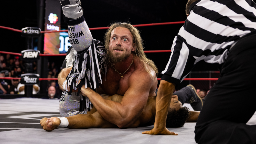 AEW wrestler Big Bill is on the mat with his leg in the air, struggling in a hold with another wrestler as the referee watches.