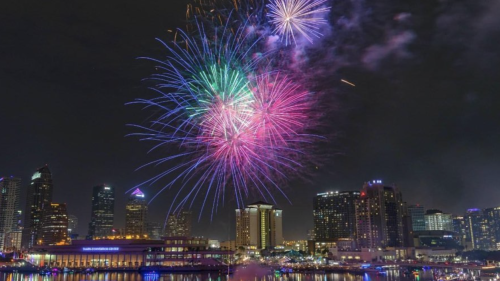 Fireworks light up the sky above downtown Tampa in 2021 during Boom of the Bay. The show is above the Hillsborough River at night time, with high rises in the background, and the Riverwalk in the foreground. The xplosions are a mix of pink, green, blue, purple and yellow.