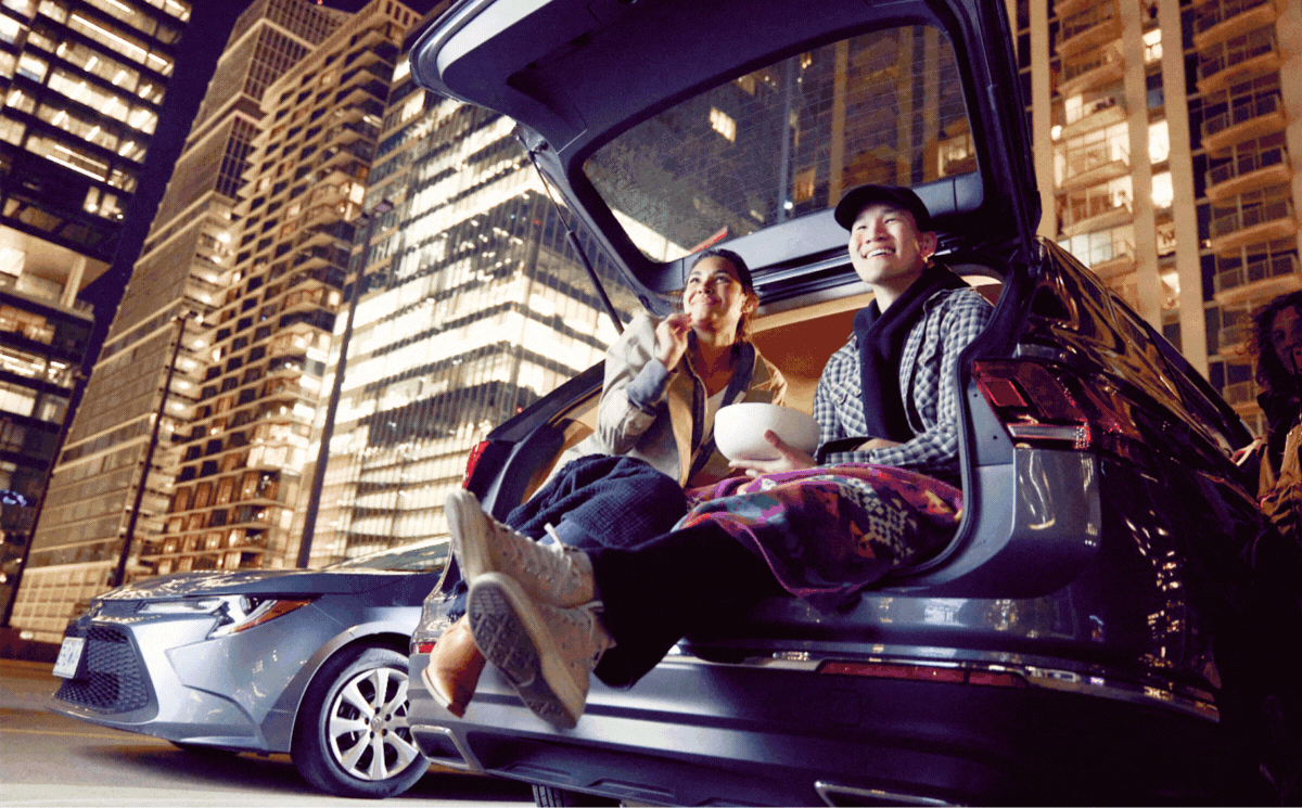 Two people sit on the back of a car, city behind them. A person gets out of a car holding a yoga mat. A person turns from the front seat and smiles.