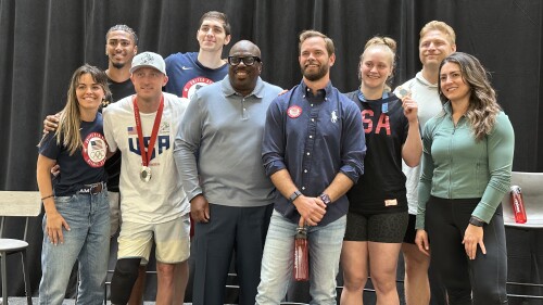 A group of US Olympians pose for a photo together for a photo on a stage.