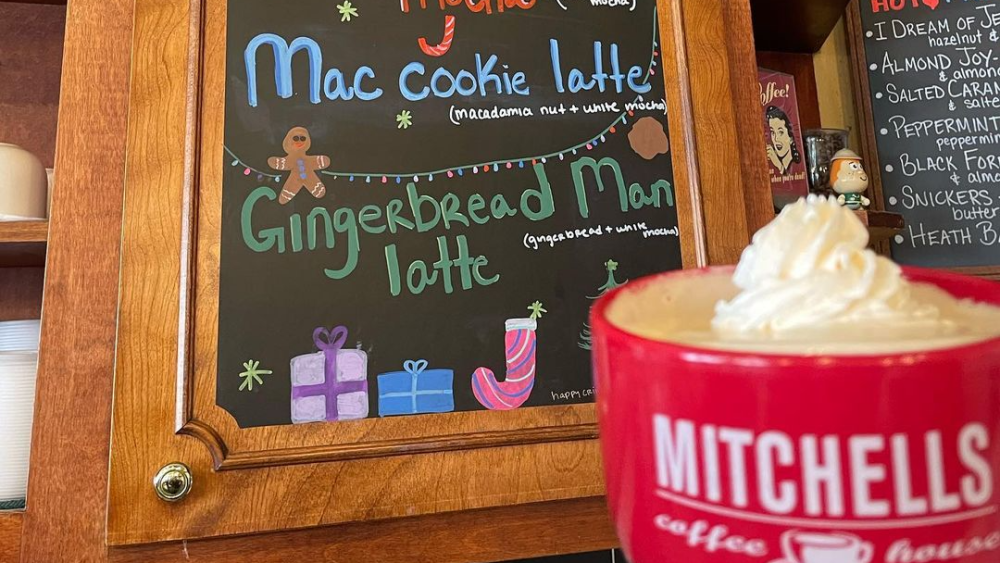 A mug of coffee with whipped cream held in front of a seasonal drink sign