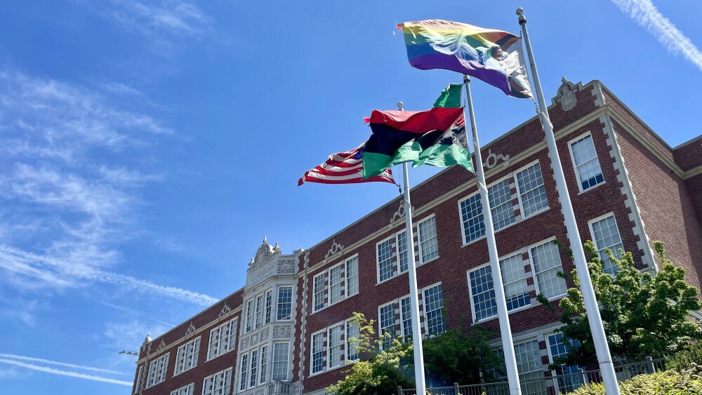 The front of historic Garfield High School features red brick and white stone, with paned glass windows and green bushes landscaped throughout the front grounds. Three flag poles hoist the American Flag, the Pan-African Flag, and the Intersectional Pride Flag high in the air between the parking lot and the building. The flags are flapping in the wind.