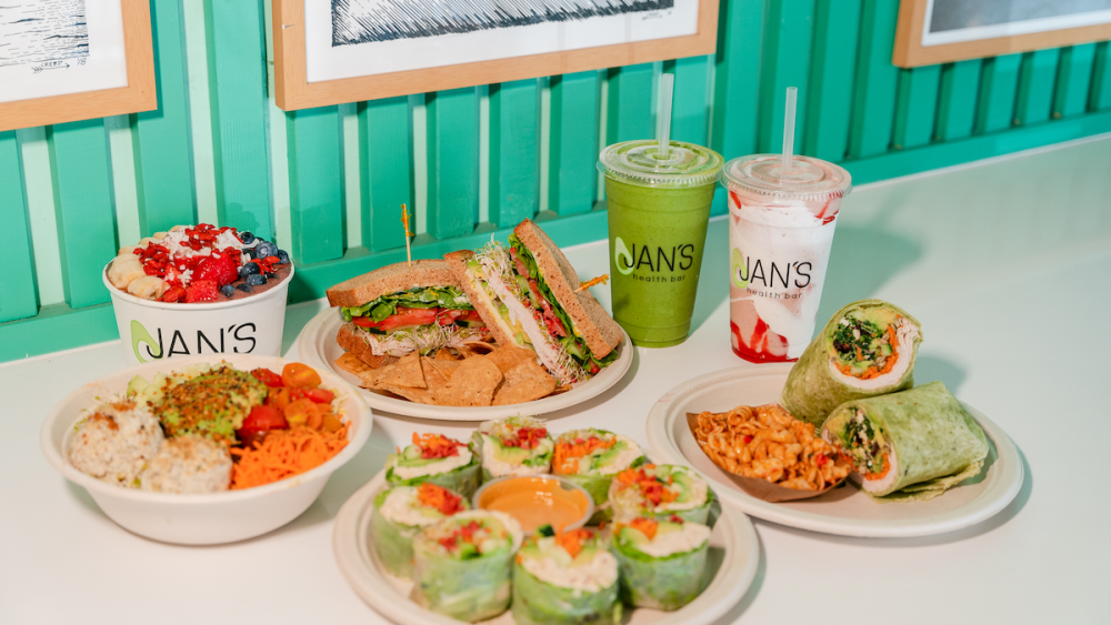A display of dishes from Jan's Health Bar including green and white smoothies, a wrap filled with turkey and vegetables, a sandwich, and an acai bowl.