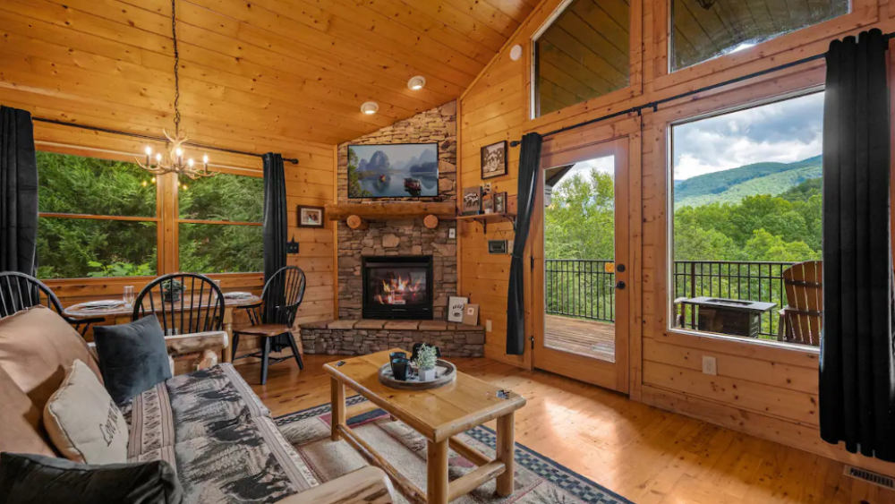 The living room of a log cabin with a stone fireplace and large windows overlooking a back deck with mountain views.
