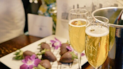 Two champagne glasses with a bucket in the background. Chocolate-covered strawberries on a white plate, roses, a card, and a print are blurred in the background.