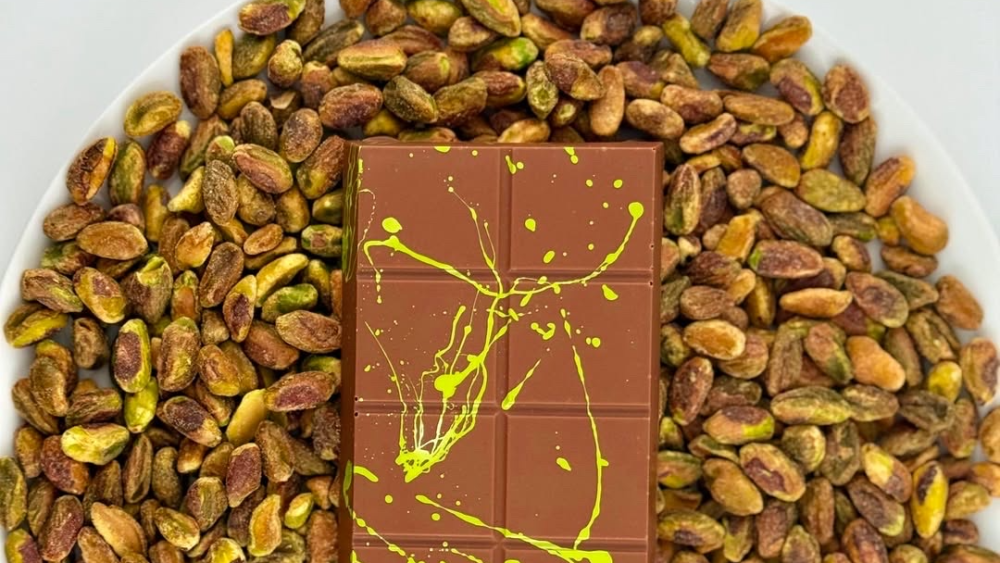 A Dubai chocolate bar with a bright green splatter design sits on top of a bed of shelled pistachio nuts.