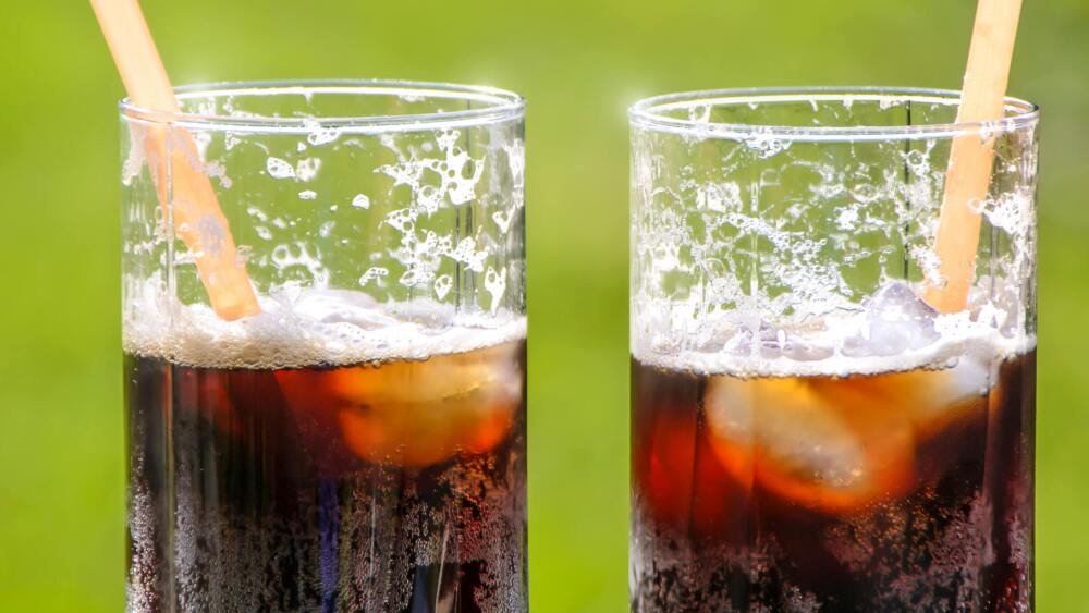 Two glasses of soda sit on a table, appearing to be filled with a cola, ice, and straws.